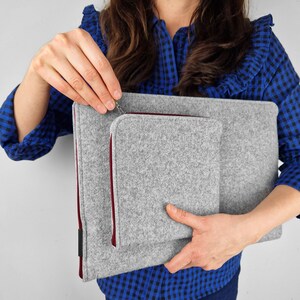 Woman holding light grey flet laptop cover. Maroon zippers. Front pocket for charger, wires or mouse. Woman opens front pocket.