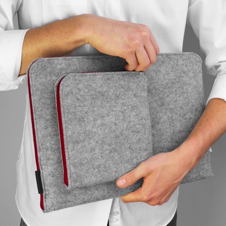 Man holding light grey flet laptop sleeve with maroon zippers and opens front pocket.