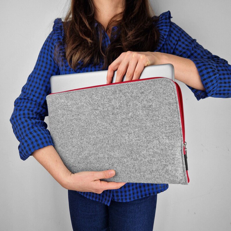 Woman holding light grey felt laptpp sleeve with red zipper. Silver laptop half out of the sleeve. Zipper open.