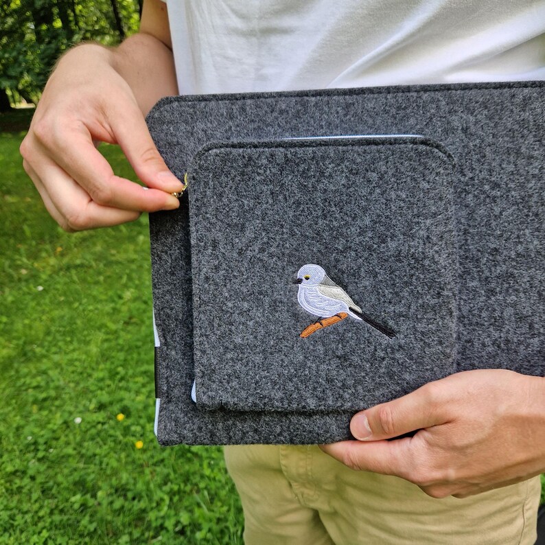Man holds dark grey felt laptop case with bird embroided on the front. Laptop sleeve has white zipper and front pocket for wires. On the pocket there is an embroidery of long-tailed tit. Cover takes 13 inch laptop.