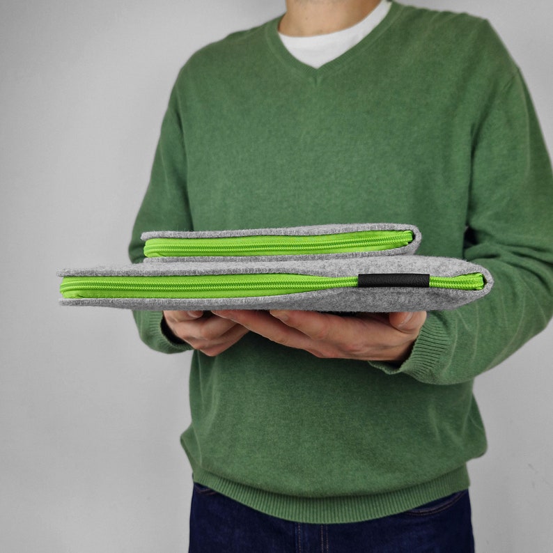 Man holding closed flet laptop sleeve flat on hands. Green zippers front to the camera. Pocket for charger sewn on the front of the sleeve.