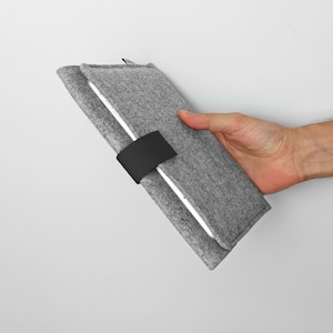 Op de afbeelding: Een grijze tablet hoes van vilt met een zwarte band sluiting. De hoes is dubbelgevouwen en wordt in een hand gehouden.