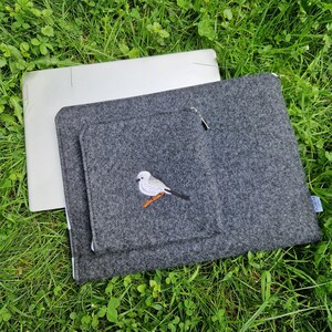 Man holds dark grey felt laptop case with bird embroided on the front. Laptop sleeve has white zipper and front pocket for wires. On the pocket there is an embroidery of long-tailed tit. Cover takes 13 inch laptop.