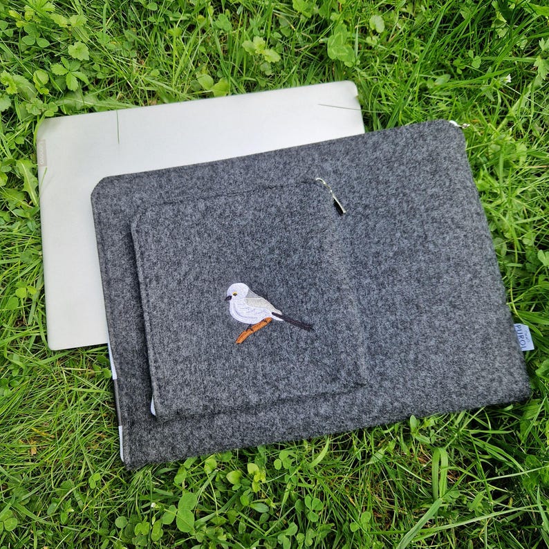 Man holds dark grey felt laptop case with bird embroided on the front. Laptop sleeve has white zipper and front pocket for wires. On the pocket there is an embroidery of long-tailed tit. Cover takes 13 inch laptop.