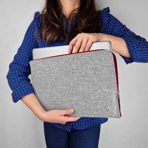 Woman hilding light grey felt laptop case with maroon zipper. Laptop half out of the sleeve.