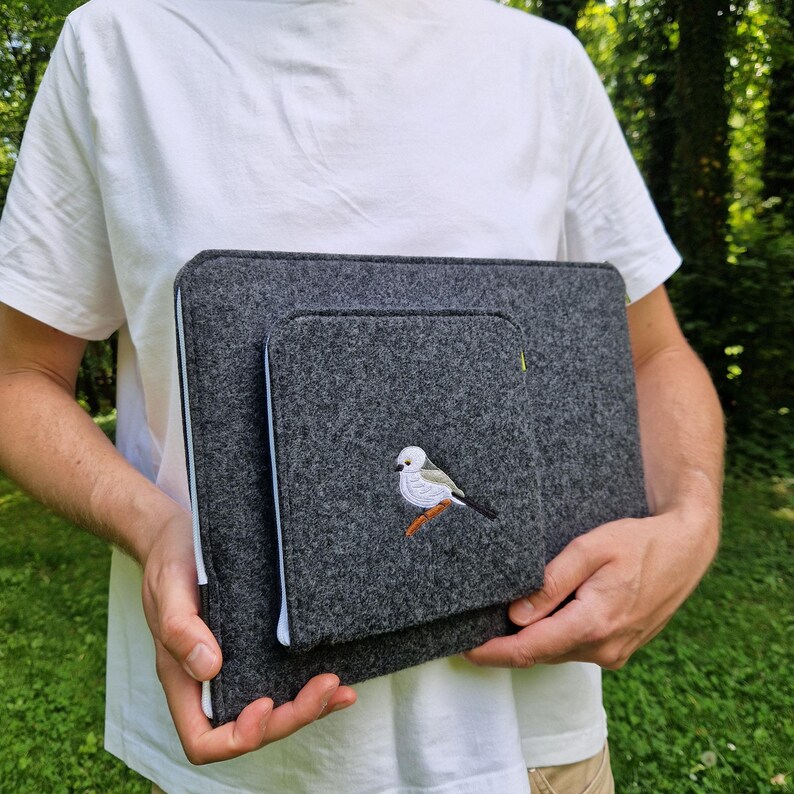 Man holds dark grey felt laptop case with bird embroided on the front. Laptop sleeve has white zipper and front pocket for wires. On the pocket there is an embroidery of long-tailed tit. Cover takes 13 inch laptop.