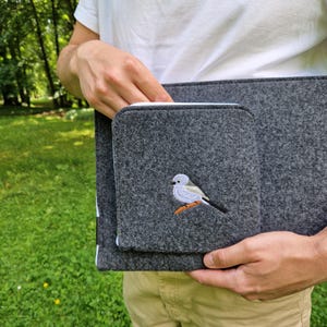 Man holds dark grey felt laptop case with bird embroided on the front. Laptop sleeve has white zipper and front pocket for wires. On the pocket there is an embroidery of long-tailed tit. Cover takes 13 inch laptop.