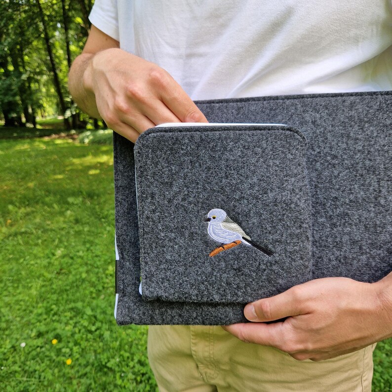 Man holds dark grey felt laptop case with bird embroided on the front. Laptop sleeve has white zipper and front pocket for wires. On the pocket there is an embroidery of long-tailed tit. Cover takes 13 inch laptop.