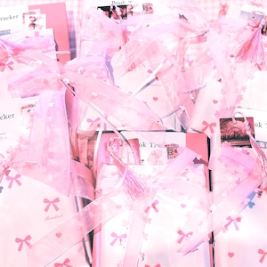 May include: Several pink gift sets, tied with sheer pink ribbons and bows. The sets contain notebooks and cards, some with the word "Tracker" printed on them. The background features a pink and white gingham pattern.