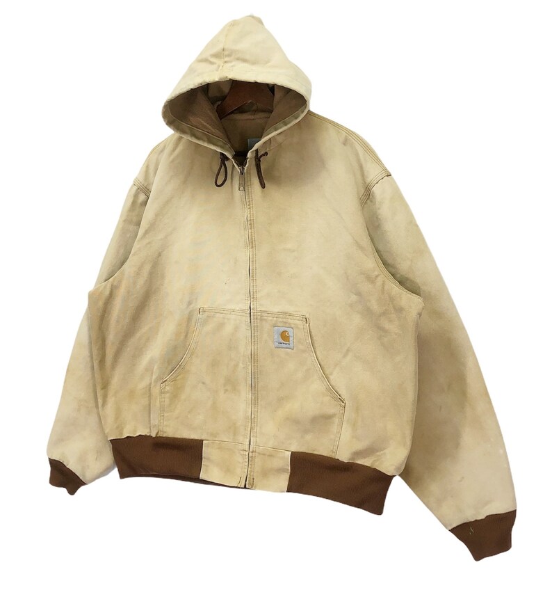 May include: A tan Carhartt hooded jacket with a brown zipper and brown trim around the bottom. The jacket has a patch on the left chest with the Carhartt logo.