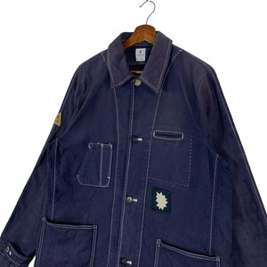 May include: A dark blue long-sleeve work jacket featuring white contrast stitching, a pointed collar, and a front button closure. The jacket includes multiple patch pockets, a small yellow circular patch on the sleeve, and a star-shaped patch.