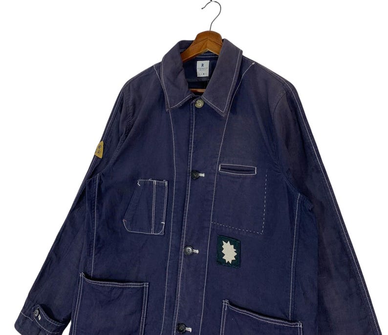 May include: A dark blue long-sleeve work jacket featuring white contrast stitching, a pointed collar, and a front button closure. The jacket includes multiple patch pockets, a small yellow circular patch on the sleeve, and a star-shaped patch.