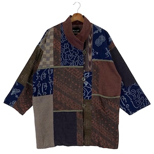 Könnte beinhalten: Patchwork-Jacke mit verschiedenen Mustern in Braun-, Blau- und Beigetönen. Die Jacke hat einen Stehkragen und Knopfverschlüsse. Das Design umfasst florale und geometrische Motive. Sie hängt an einem Holzkleiderbügel.