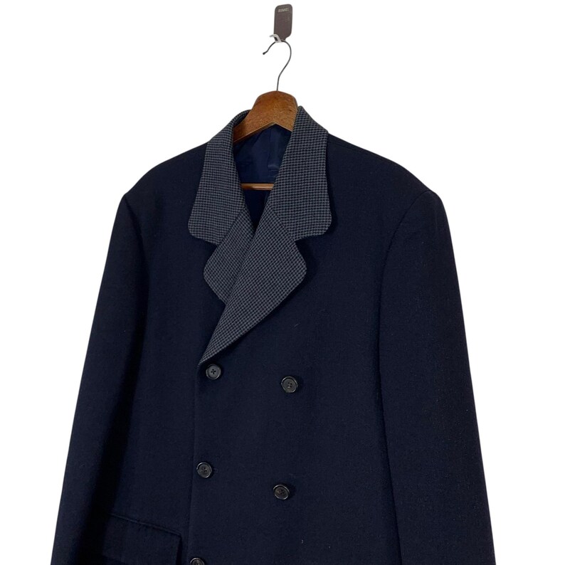 Puede incluir: Un abrigo azul marino de doble botonadura con un cuello estampado gris y blanco en contraste. El abrigo tiene botones negros y est&aacute; colgado de una percha de madera. El abrigo es de estilo cl&aacute;sico.