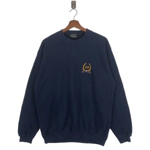 Può includere: Una felpa blu navy con un logo ricamato in oro che presenta le lettere "CD" all'interno di una corona. Il logo si trova sul petto sinistro della felpa.