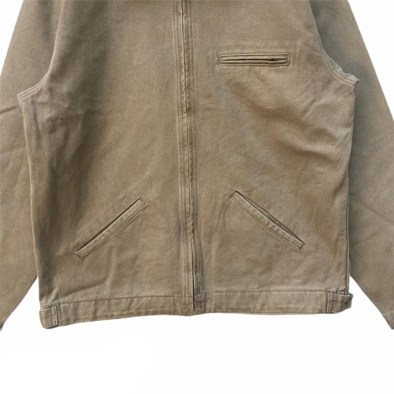 K&ouml;nnte beinhalten: Eine braune Canvas-Jacke mit Rei&szlig;verschluss und zwei Taschen auf der Vorderseite. Die Jacke hat eine lockere Passform und ein leicht verblichenes Aussehen.