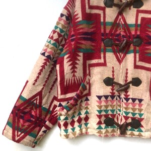 May include: A cropped jacket with a Southwestern-inspired geometric pattern in red, beige, teal, and purple. The jacket features toggle closures and a pocket. The sleeve and body are visible, showcasing the detailed design.