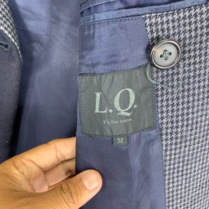 Puede incluir: Primer plano del interior de una blazer azul marino con estampado de pata de gallo. La blazer presenta un bot&oacute;n y una etiqueta que dice "L.Q. Y's for men" con una talla "M". El forro es de color azul marino liso.