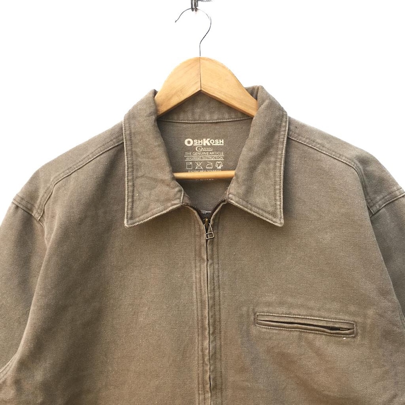 K&ouml;nnte beinhalten: Eine braune OshKosh B'Gosh Arbeitsjacke mit Rei&szlig;verschluss und einer einzigen Tasche auf der linken Seite. Das Etikett lautet "OshKosh B'Gosh The Genuine Article Made in the USA."