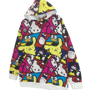 Op de afbeelding: Een wit hoodie met een kleurrijke all-over print met cartoonfiguren zoals Hello Kitty, My Melody en Keroppi. De print is een mix van roze, geel, blauw, rood en zwart.