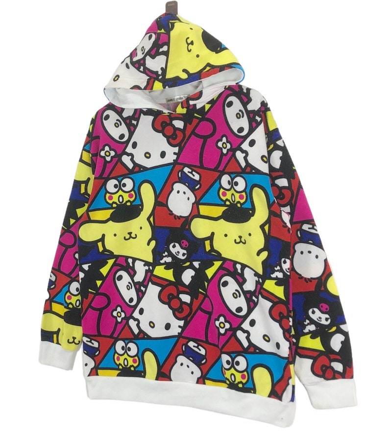 Op de afbeelding: Een wit hoodie met een kleurrijke all-over print met cartoonfiguren zoals Hello Kitty, My Melody en Keroppi. De print is een mix van roze, geel, blauw, rood en zwart.