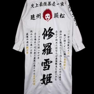 Pode incluir: Camisa de manga comprida de cor clara, com texto japonês e um gráfico vermelho e preto nas costas. A camisa tem uma gola clássica e punhos abotoados, com texto também nas mangas.