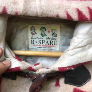 May include: Close-up of a beige and red patterned jacket with a wooden toggle closure. The jacket features a label with the text "From Pupils to Workers B &raquo; SPARE High Spirits, Brightly, Healthy." The jacket is hanging on a wooden hanger.