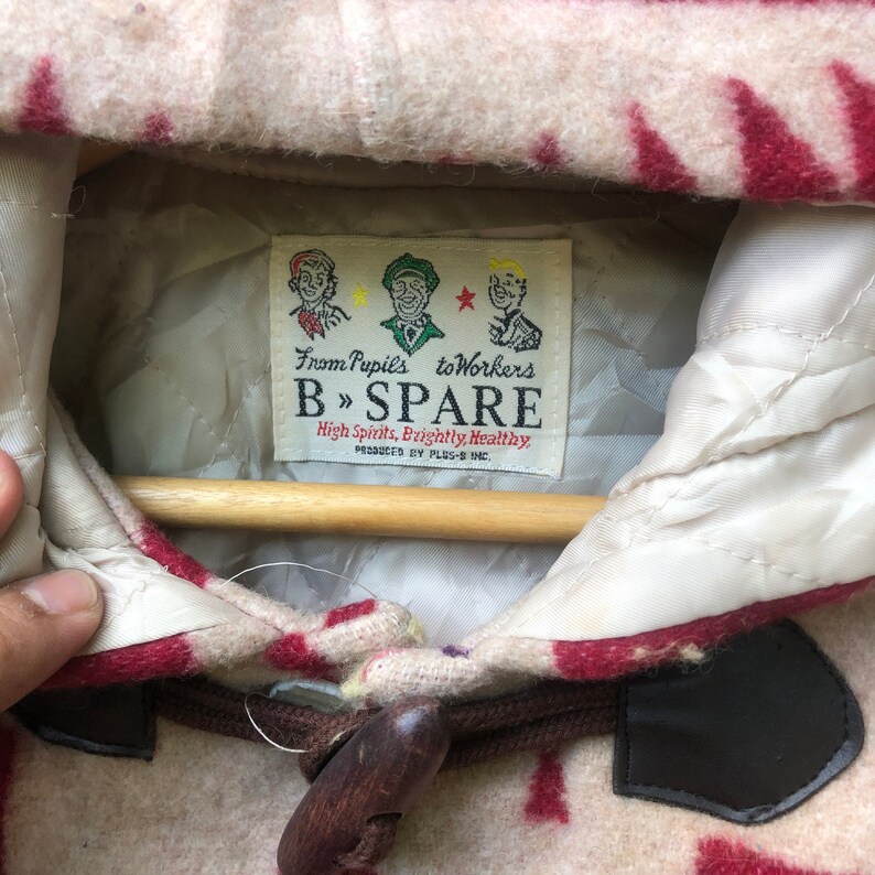 May include: Close-up of a beige and red patterned jacket with a wooden toggle closure. The jacket features a label with the text "From Pupils to Workers B &raquo; SPARE High Spirits, Brightly, Healthy." The jacket is hanging on a wooden hanger.