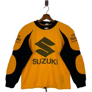 May include: A yellow sweatshirt with black and yellow shoulder pads. The sweatshirt has a large green Suzuki logo and the text "SUZUKI" in green letters. The text "ITALY SUZUKI RACING" is on the right shoulder and the text "ITALY SUZUKI TEAM" is on the left shoulder.