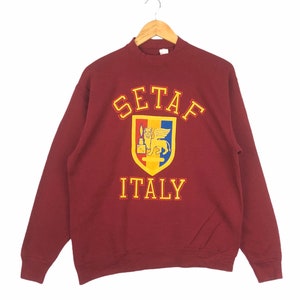 May include: A maroon sweatshirt with a yellow and white graphic of a crest with a lion and the text "SETAF ITALY".