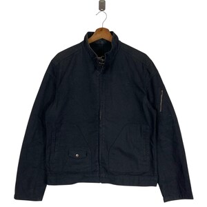 May include: Black jacket with a stand-up collar, a zip closure, and multiple pockets. The jacket is hanging on a wooden hanger against a white background. The jacket has a small pocket on the left chest and a zip pocket on the left sleeve.