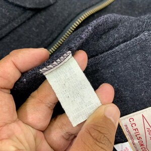 Puede incluir: Primer plano de una prenda de lana gris oscuro con cremallera. Se ve una etiqueta blanca con texto negro, que enumera detalles como "SEATTLE, WA" y "100% VIRGIN WOOL".
