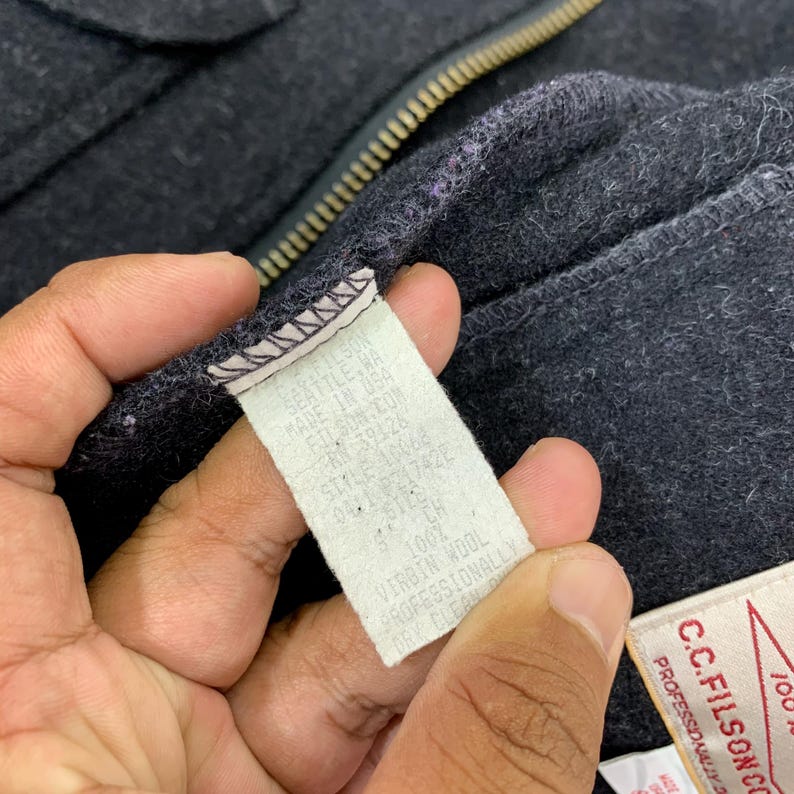 Puede incluir: Primer plano de una prenda de lana gris oscuro con cremallera. Se ve una etiqueta blanca con texto negro, que enumera detalles como "SEATTLE, WA" y "100% VIRGIN WOOL".