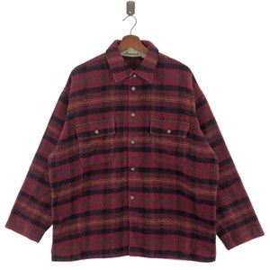 May include: A red, black, and brown checked shirt with two chest pockets. The shirt is suspended from a wooden hanger. The collar label reads "Almanack".