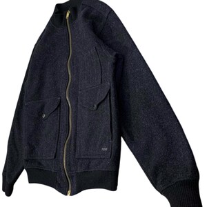 Puede incluir: Chaqueta bomber de lana gris oscuro con cremallera de lat&oacute;n de largo completo y dos bolsillos delanteros. La chaqueta tiene pu&ntilde;os y cuello de canal&eacute; negros. La marca "FILSON" es visible en el bolsillo derecho.