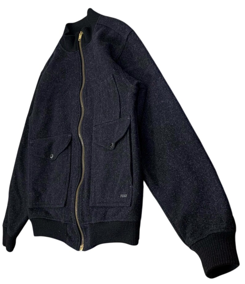 Puede incluir: Chaqueta bomber de lana gris oscuro con cremallera de lat&oacute;n de largo completo y dos bolsillos delanteros. La chaqueta tiene pu&ntilde;os y cuello de canal&eacute; negros. La marca "FILSON" es visible en el bolsillo derecho.