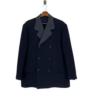 Puede incluir: Un abrigo azul marino de doble botonadura con un cuello estampado gris y blanco en contraste. El abrigo tiene dos bolsillos delanteros y seis botones. El abrigo est&aacute; colgado de una percha de madera.