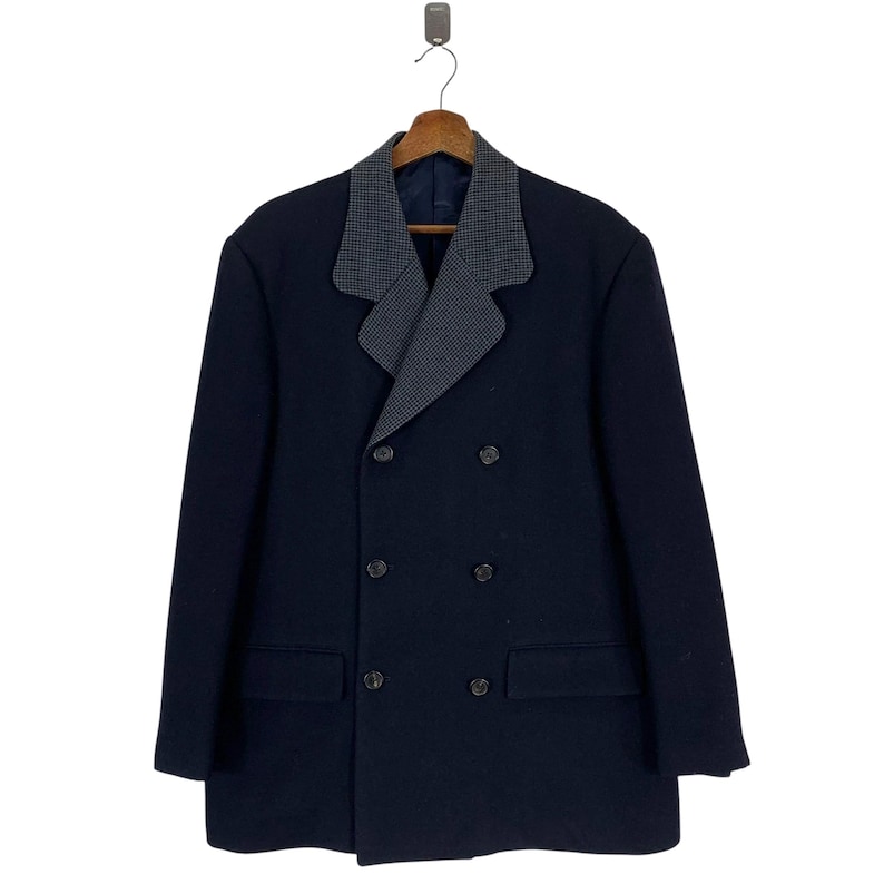 Puede incluir: Un abrigo azul marino de doble botonadura con un cuello estampado gris y blanco en contraste. El abrigo tiene dos bolsillos delanteros y seis botones. El abrigo est&aacute; colgado de una percha de madera.