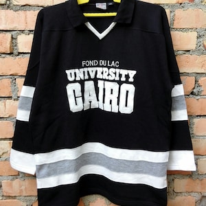 May include: Black long-sleeve shirt with white and gray stripes. The shirt has a polo collar and features the text "Fond du Lac University Cairo" in white.
