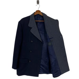 Puede incluir: Un abrigo cruzado azul marino con solapa y forro estampados en gris y blanco contrastantes. El abrigo est&aacute; colgado de una percha de madera. El forro interior es azul marino liso. El abrigo tiene dos filas de botones y un bolsillo a cada lado.