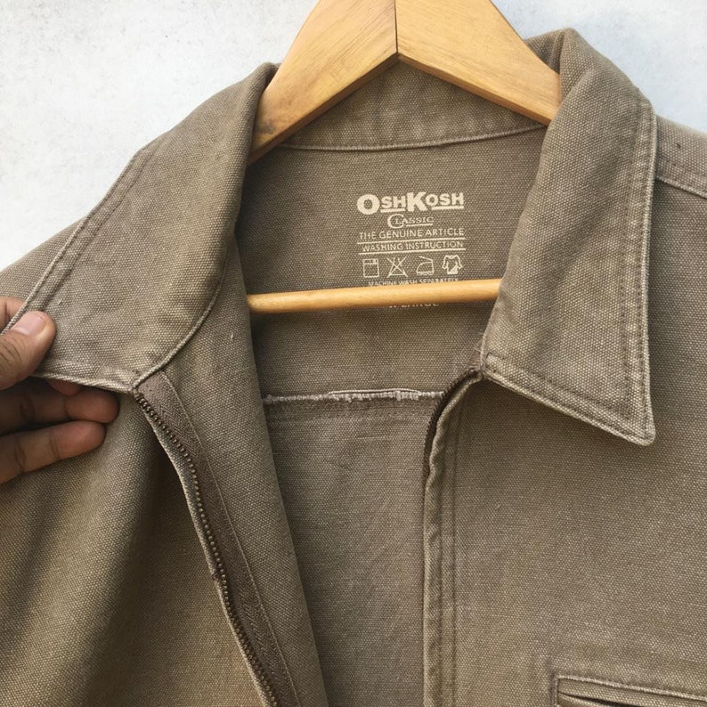 K&ouml;nnte beinhalten: Eine braune OshKosh B'Gosh Classic Zip-Jacke mit einem Etikett mit der Aufschrift "The Genuine Article" und Waschhinweisen.