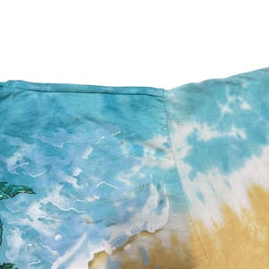 May include: A tie-dye t-shirt featuring a beach scene with a blue ocean, white waves, and a yellow and tan sandy shore. The shirt has a crew neck and short sleeves. The design includes a green palm tree.