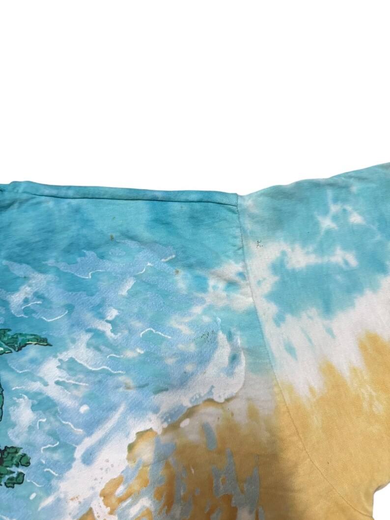 May include: A tie-dye t-shirt featuring a beach scene with a blue ocean, white waves, and a yellow and tan sandy shore. The shirt has a crew neck and short sleeves. The design includes a green palm tree.
