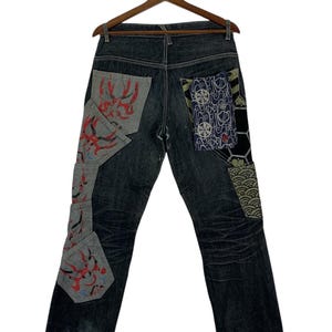 Può includere: Jeans in denim blu scuro con dettagli patchwork. I jeans presentano toppe con disegni astratti rossi e neri e motivi di ispirazione giapponese. I jeans sono appesi a una gruccia di legno.