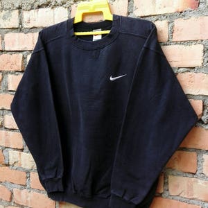 May include: A black Nike sweatshirt with a white Swoosh logo on the front. The sweatshirt is hanging on a yellow hanger against a brick wall.