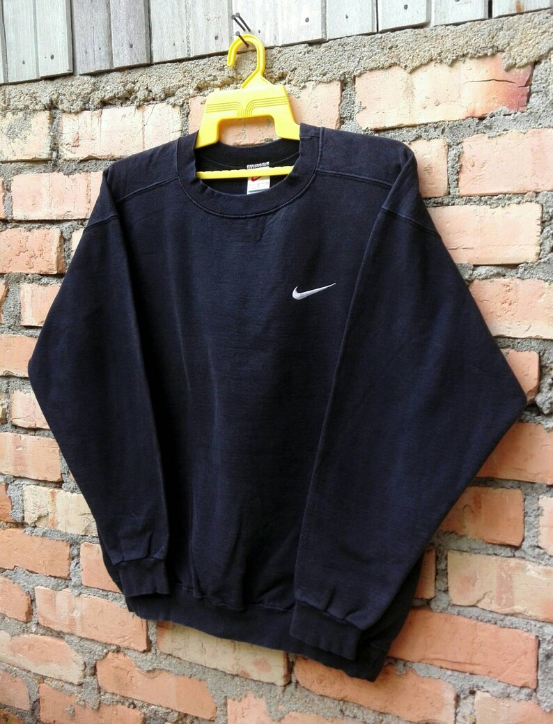 May include: A black Nike sweatshirt with a white Swoosh logo on the front. The sweatshirt is hanging on a yellow hanger against a brick wall.