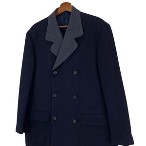 Puede incluir: Un abrigo azul marino de doble botonadura con un cuello estampado gris y blanco en contraste. El abrigo tiene seis botones y dos bolsillos delanteros. Est&aacute; colgado de una percha de madera.