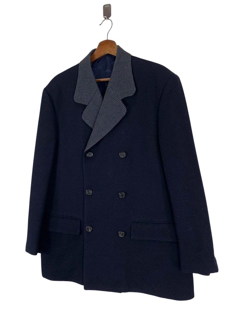 Puede incluir: Un abrigo azul marino de doble botonadura con un cuello estampado gris y blanco en contraste. El abrigo tiene seis botones y dos bolsillos delanteros. Est&aacute; colgado de una percha de madera.