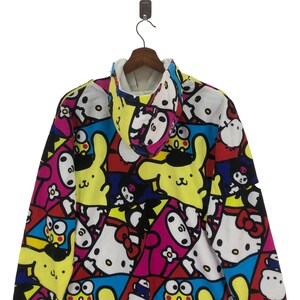 Op de afbeelding: Een kleurrijk hoodie met een veelkleurige print met cartoonfiguren. De print bevat een gele konijn, een roze kat, een blauwe kikker en een zwart-wit personage met een schedel en gekruiste beenderen.