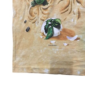 May include: A tan t-shirt featuring a detailed graphic of sea turtles. One turtle is emerging from an egg, while another is depicted above. The design includes sand-like textures and the text "&copy;1996 LIQUID BLUE".